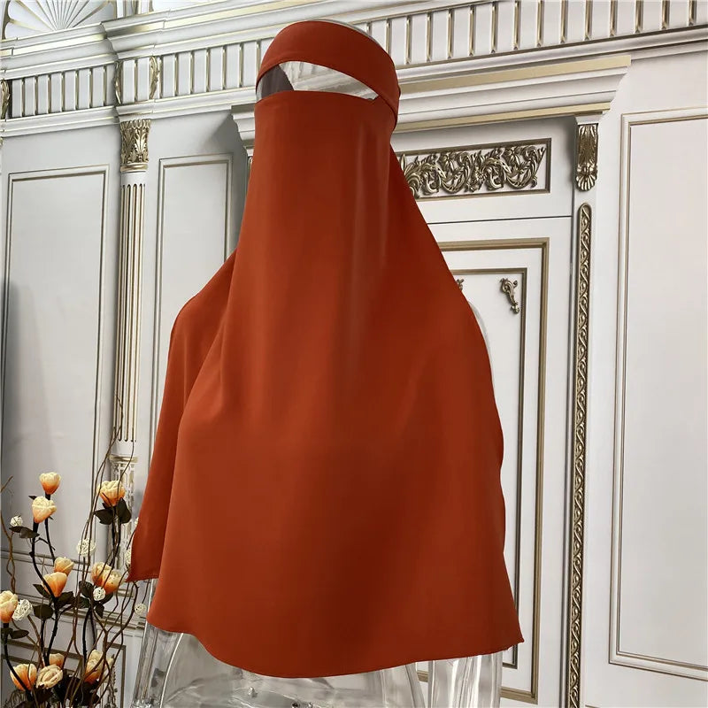Fließendes, rostfarbenes Maxi-Abaya auf einem klaren Gestell vor einem verzierten weißen und goldenen Türrahmen.