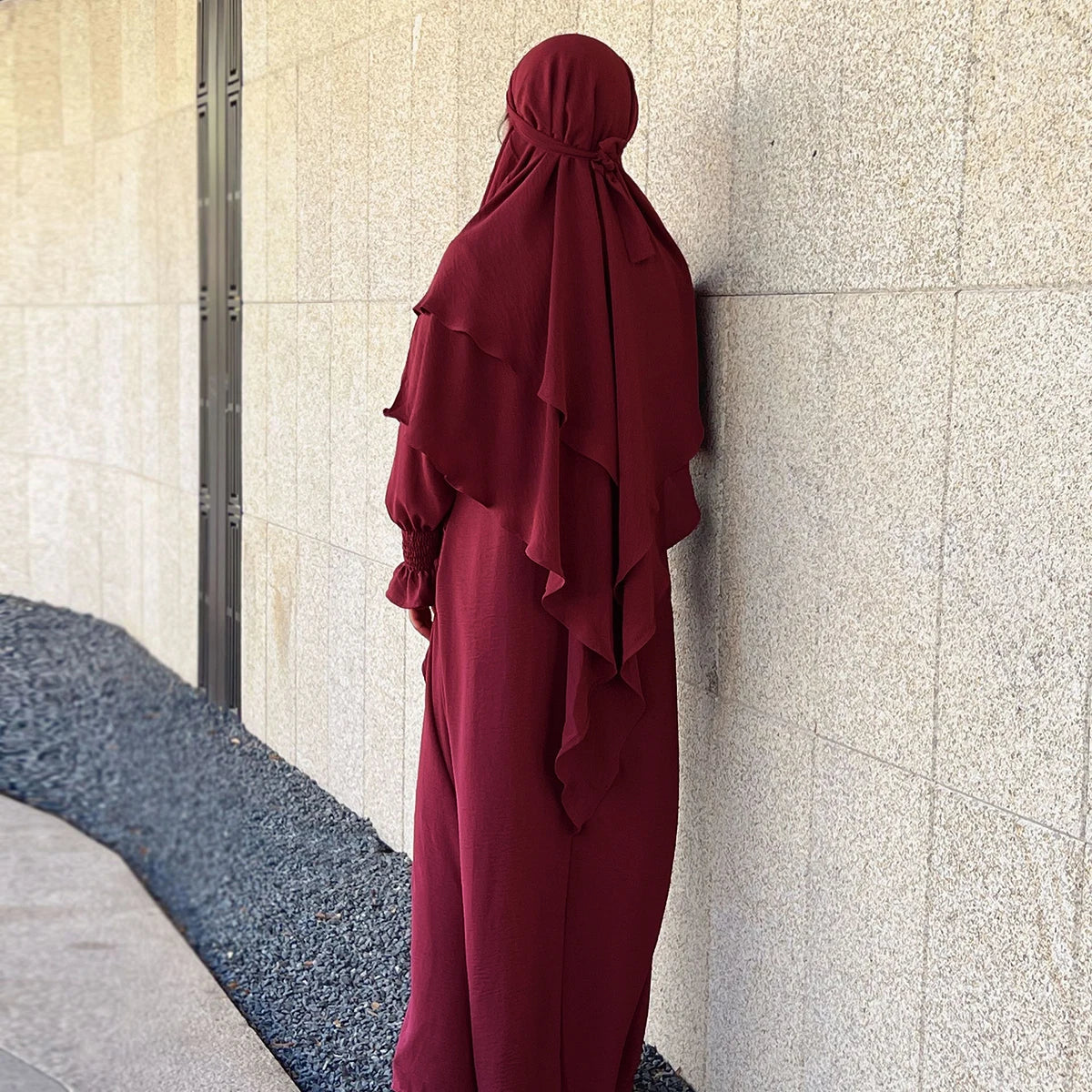 A woman wearing a maroon abaya and khimar set standing against a light-colored wall.