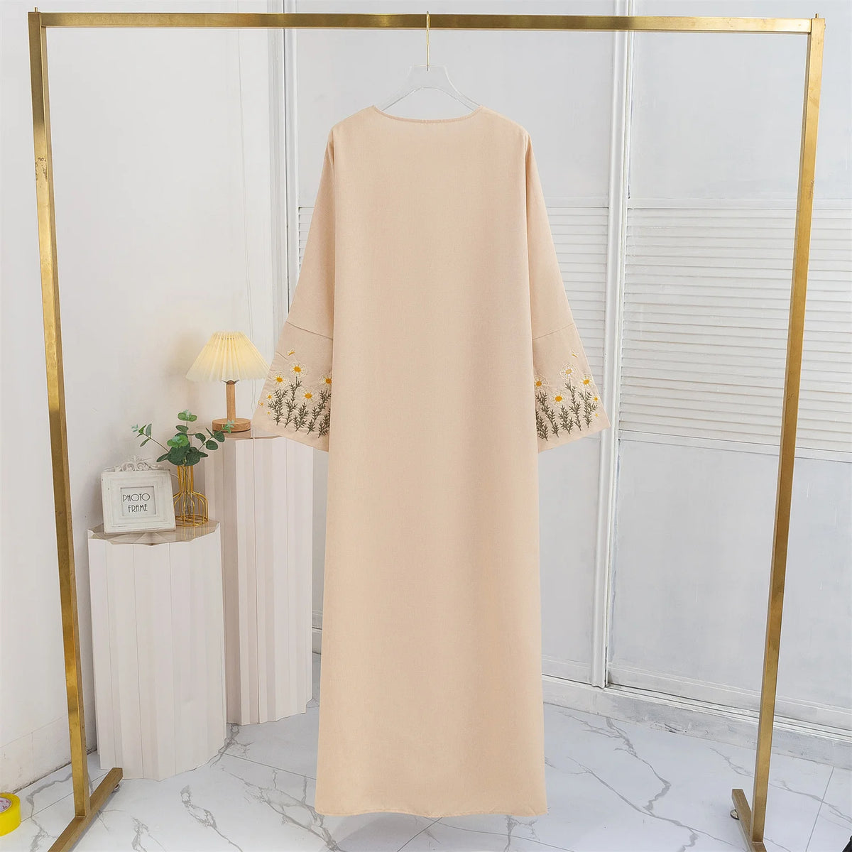 Beige Maxi-Abaya mit Blumenstickerei auf goldener Kleiderstange in einem hellen Raum.