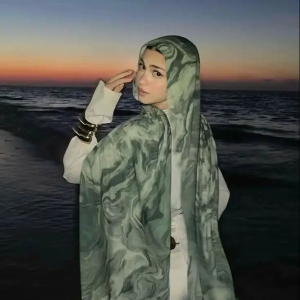 A woman wearing an elegant printed cotton rayon hijab scarf with a light green pattern, looking towards the sea at sunset.