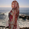 A woman wearing an elegant watercolor print modal scarf in shades of pink and brown, looking out over the ocean.