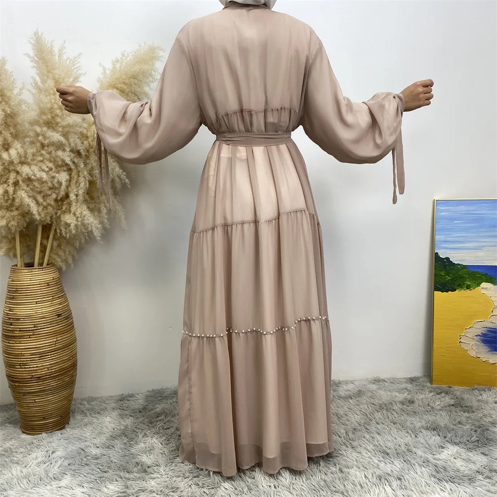 Abrigo cardigan de abaya con cuentas beige, mostrado contra una pared blanca con hierbas secas.