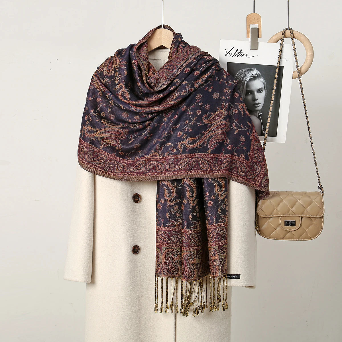 Chal de bordado paisley morado oscuro y granate sobre un abrigo color crema, con un bolso de cuero cerca.