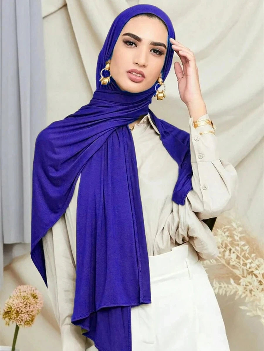 Una mujer vistiendo un pañuelo tipo jersey morado vibrante y una blusa beige clara con pantalones blancos.