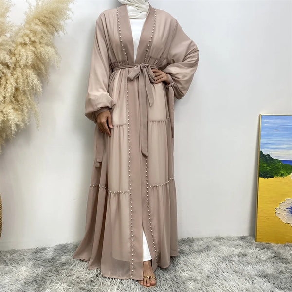 A model wearing a beige beaded abaya cardigan robe with a white top, standing against a white wall.