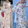 Una mujer modela un vestido kaftan maxi de estampado floral en tonos rosa claro y blanco contra un fondo de mar azul.