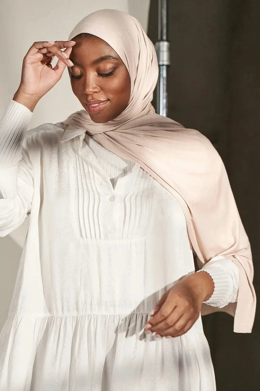 Una mujer vistiendo un pañuelo de hijab de jersey modal beige premium y una blusa blanca de manga larga.