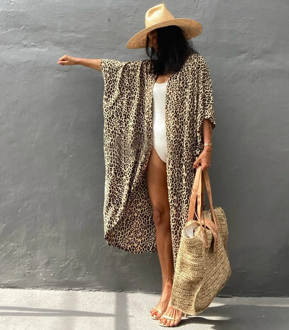 Eine Frau mit Leopardenmuster-Kimono-Überwurf über einem weißen Kleid und einem Strohhut, vor einer grauen Wand posiert.