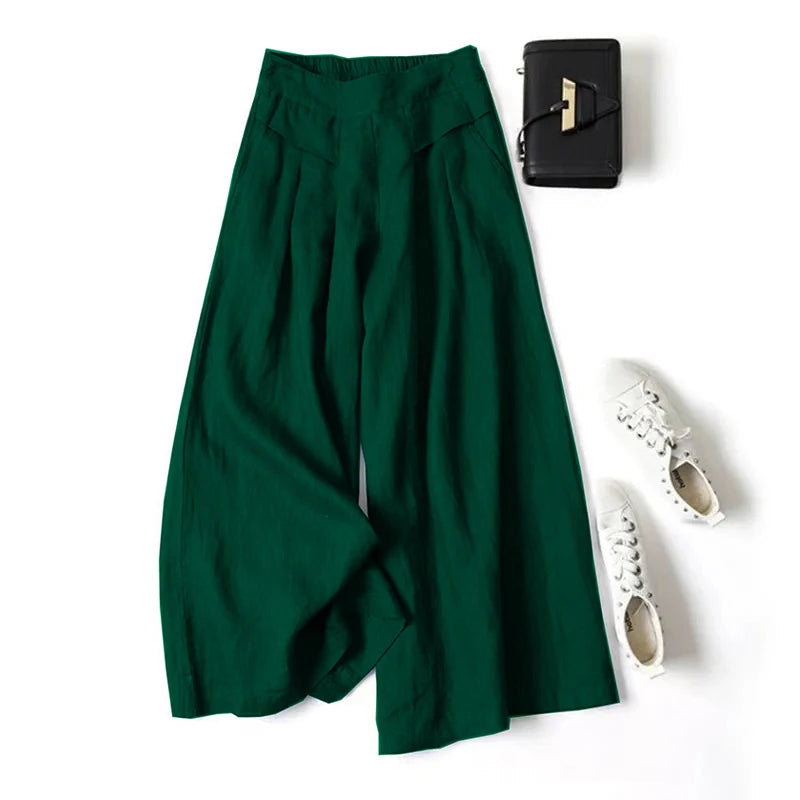 Ein Paar dunkelgrüne Leinenmisch-Palazzo-Hosen mit Cropped-Schnitt, flach auf einer schwarzen Ledergeldbörse und weißem...