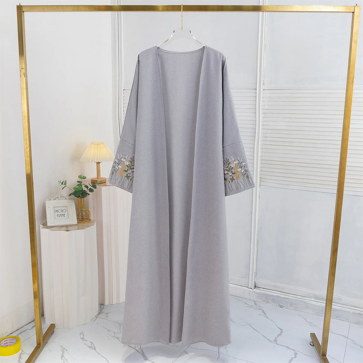 Lange graue Abaya mit Blumenstickerei auf goldener Kleiderbügel in einem hellen Raum.