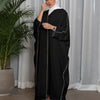 Una mujer vistiendo una abaya negra oversize con ribetes blancos y un hijab blanco, de pie frente a una cortina transparente.
