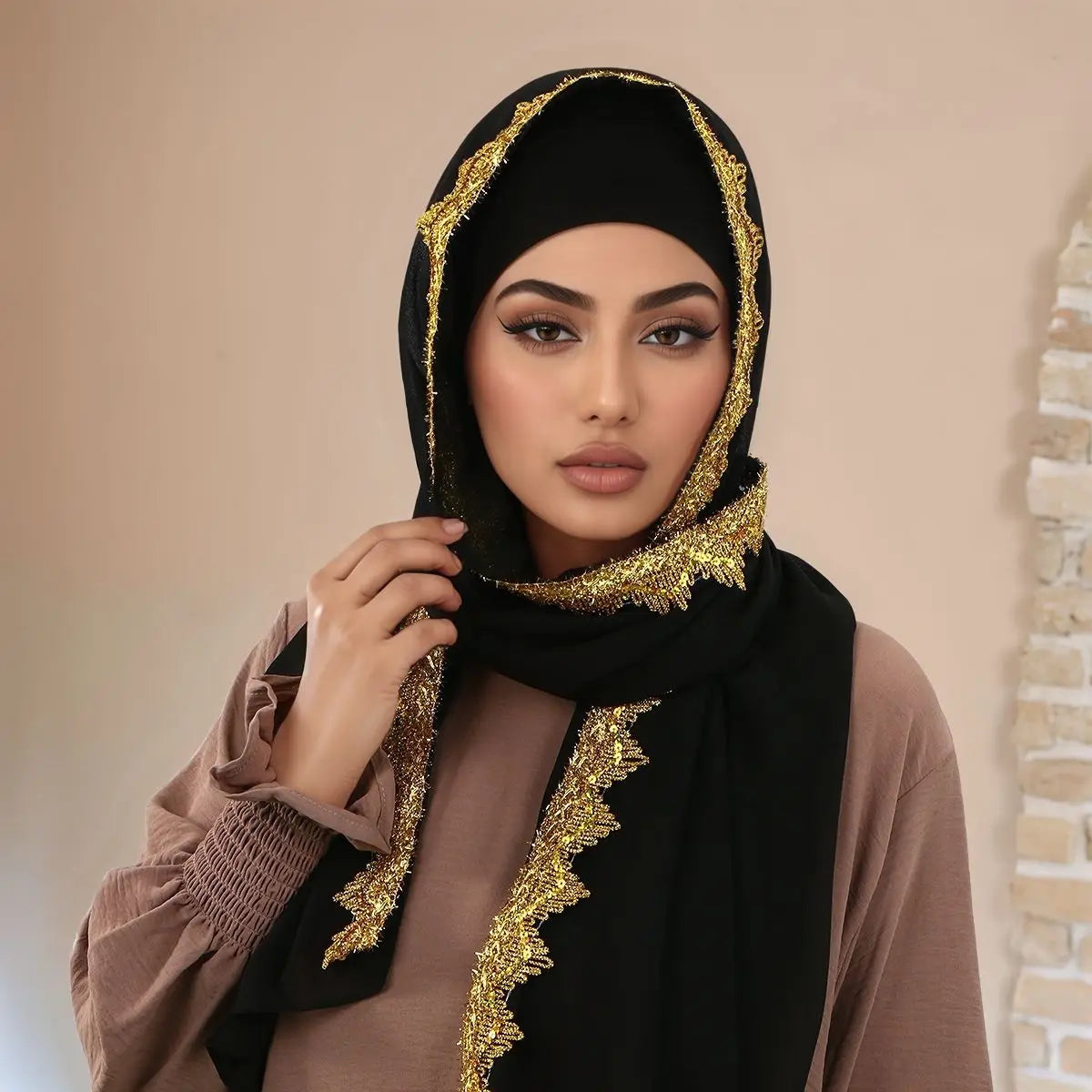 Eine Frau trägt einen schwarzen Spitzen-Chiffon-Hijab mit goldener Stickerei und einem beigen Oberteil.