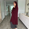Eine Frau in einem dunkelroten Abaya und Hijab steht in einem Flur mit einem weißen Sofa.