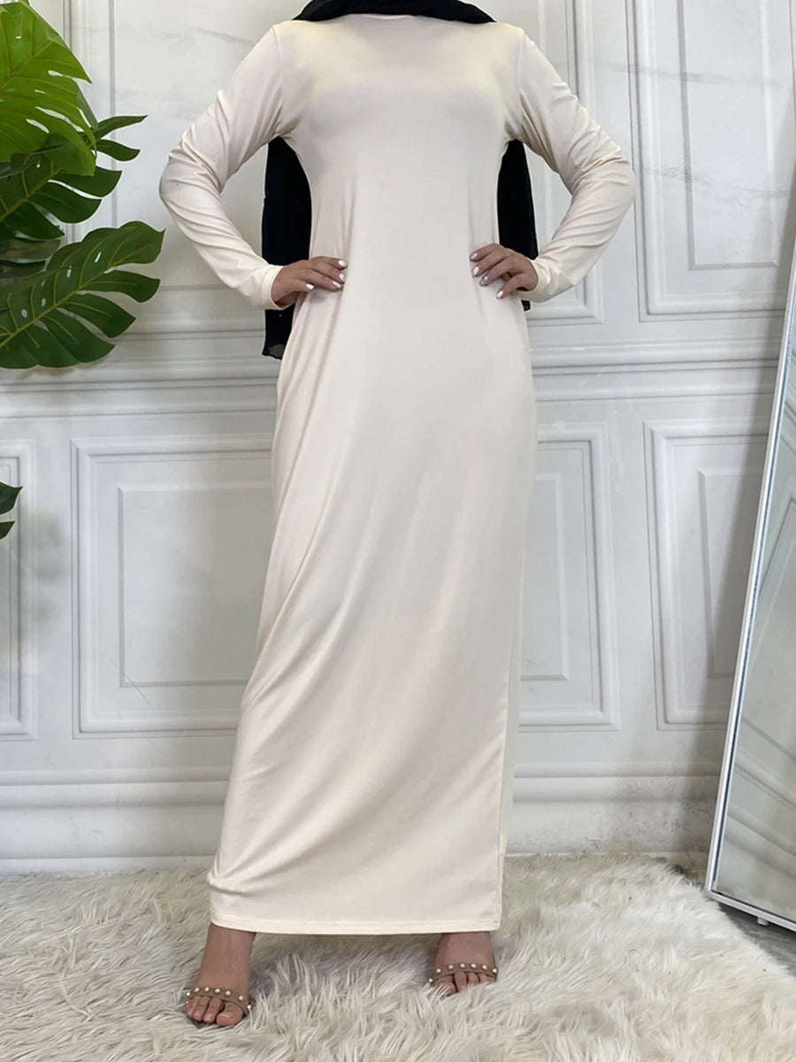 Eine Frau in einem eleganten beigen Maxi-Abaya-Kleid mit schwarzem Hijab vor einer weißen Paneelwand.