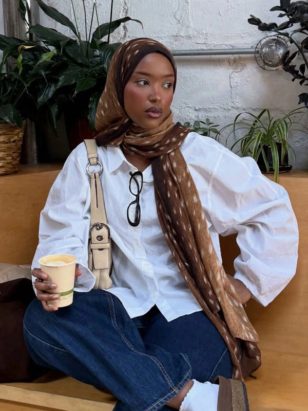 A woman wearing a white shirt and blue jeans with a brown watercolor deer print hijab scarf.