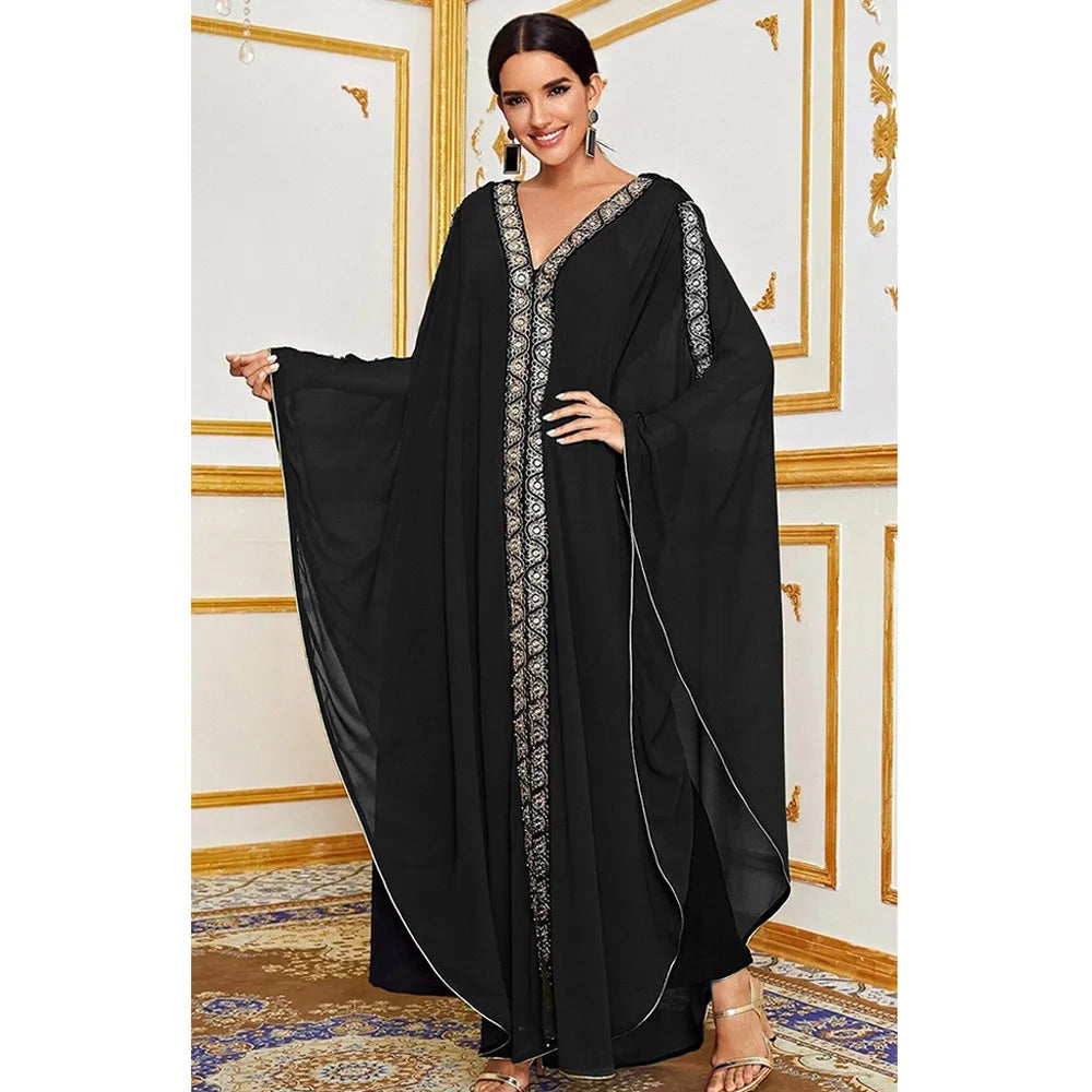 Una mujer modela un vestido maxi abaya negro y fluído con mangas de murciélago y bordado dorado contra un fondo claro.