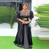 A woman in a long black sequin African party dress stands outdoors with lush green foliage.