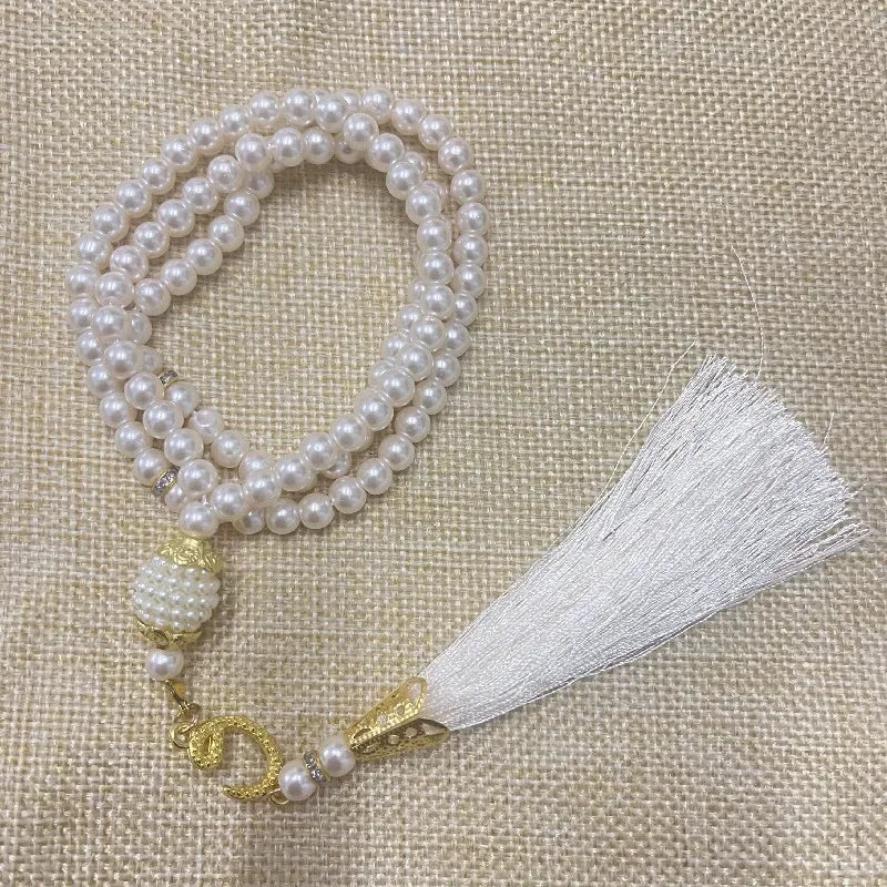 Weiße Perlenrosary-Armband mit Quaste und Goldakzenten auf beigem Stoff.