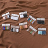 A collection of 8 solid color brooch pins in various shades are displayed in small clear plastic pouches on a brown fabric.