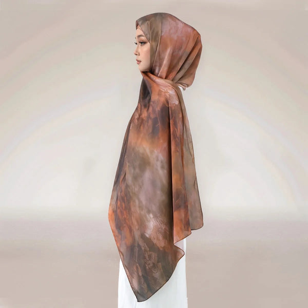 A woman wearing an elegant printed silk hijab shawl in a warm brown tone.
