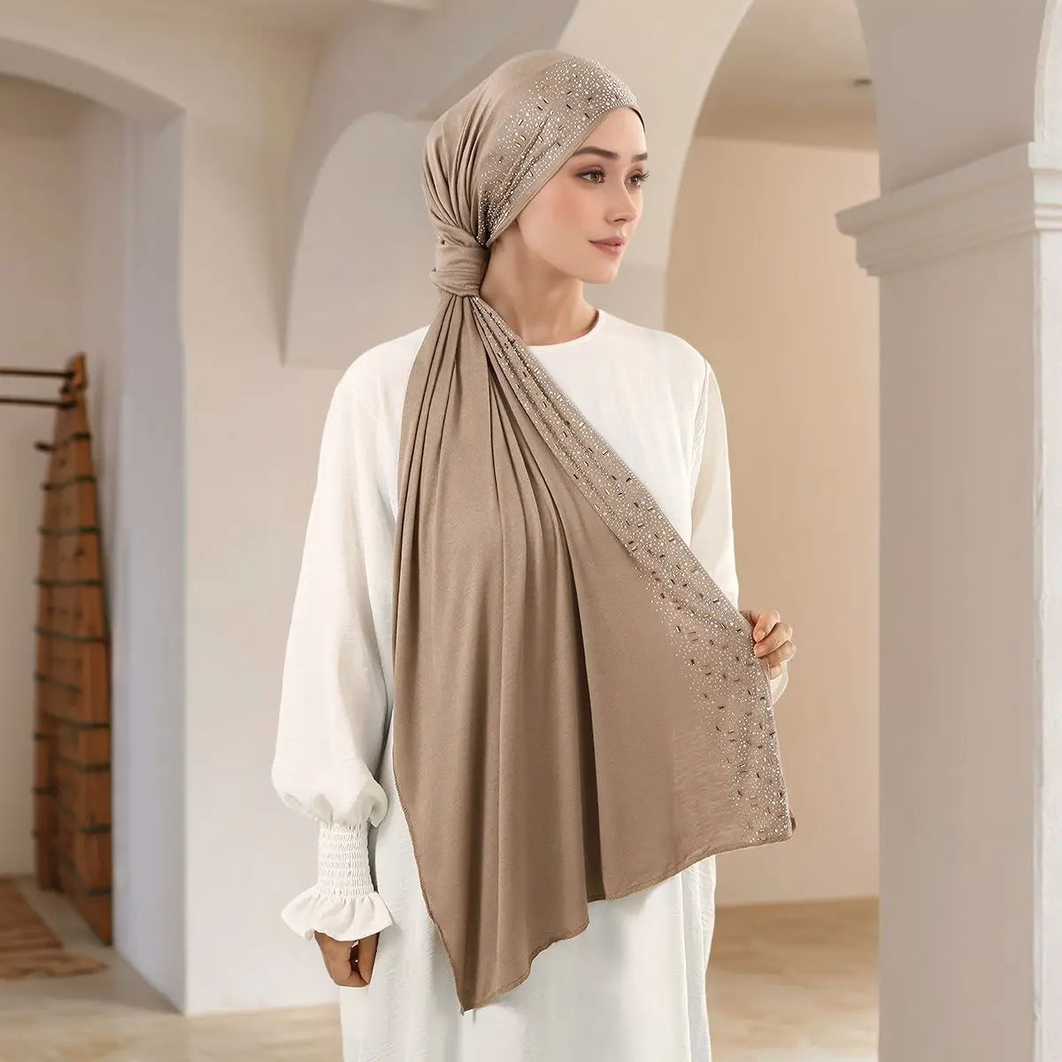 Eine Frau trägt einen beigen Diamant-Schal in Turban-Stil, drapiert über einem weißen Oberteil, in einem Innenraum.