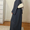 Une femme portant une abaya ample de couleur unie et un hijab gris clair se tient dans une pièce bien éclairée.