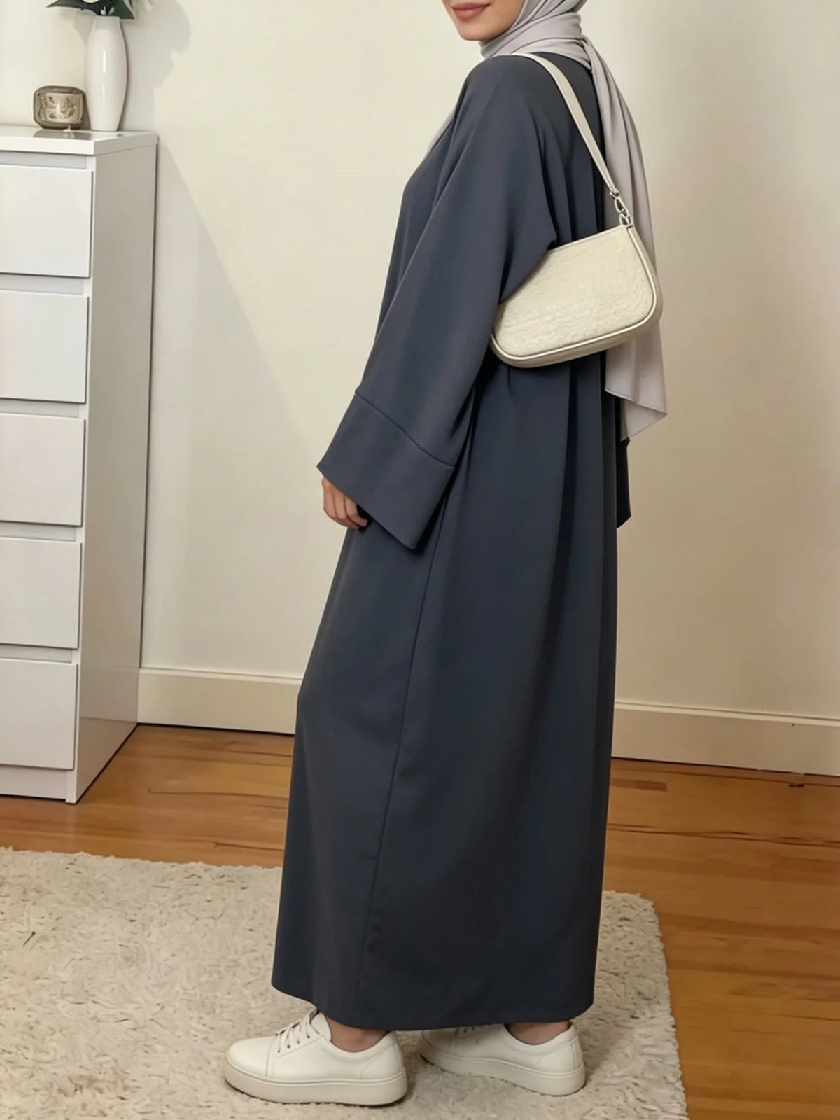 Une femme portant une abaya ample de couleur unie et un hijab gris clair se tient dans une pièce bien éclairée.