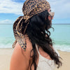 A woman with long dark hair wearing a leopard print silk satin neckerchief hijab scarf stands near the ocean.