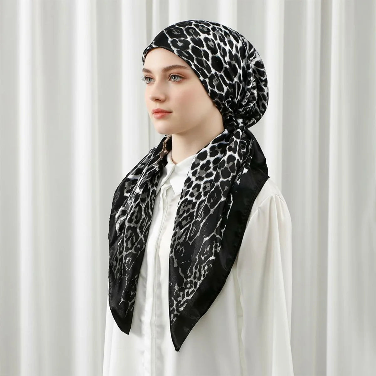 Une femme portant un foulard carré à motif léopard noir et un haut blanc sur fond clair.