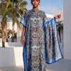 Eine Frau trägt ein elegantes bedrucktes Seidenkaftankleid in blau-goldenen Mustern im Freien.