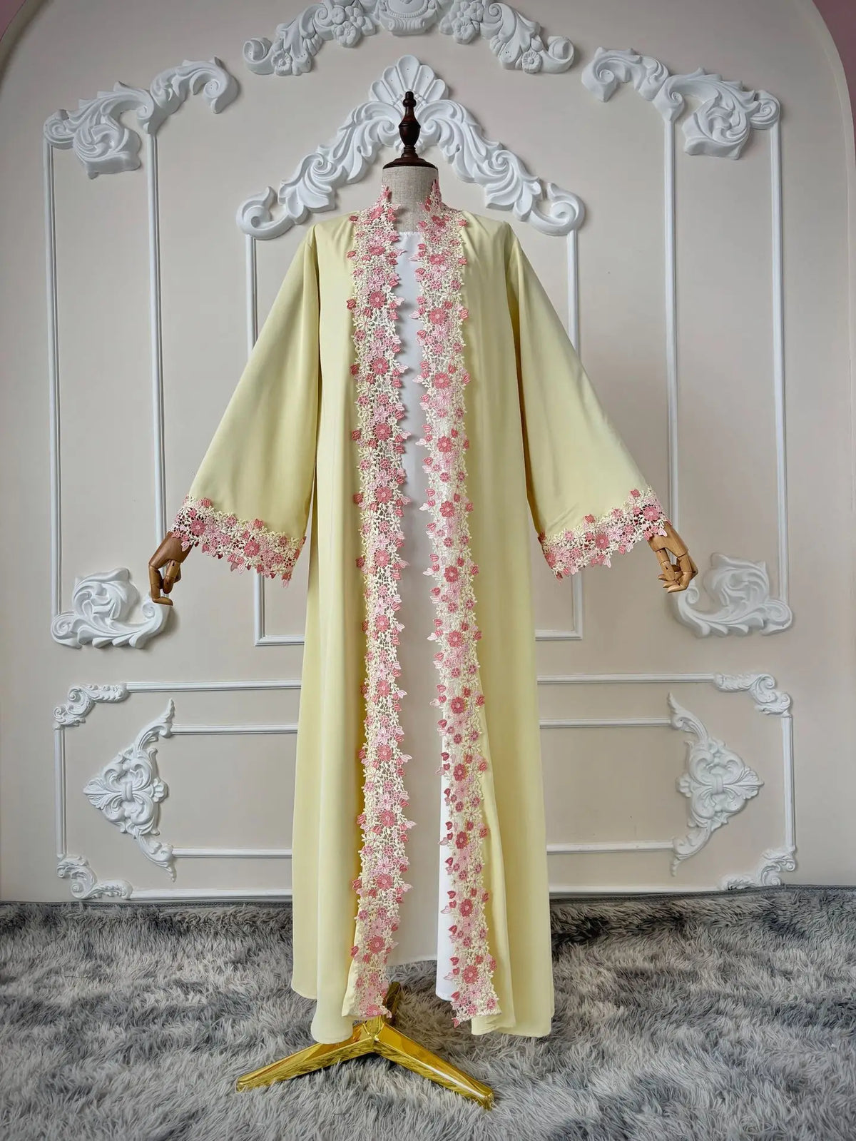 Une longue abaya jaune pâle avec bordure de dentelle florale rose, présentée sur un mannequin devant un fond arqué blanc.