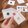 Una mano sostiene y organiza imanes de hijab de varios colores sobre una superficie de tela marrón.