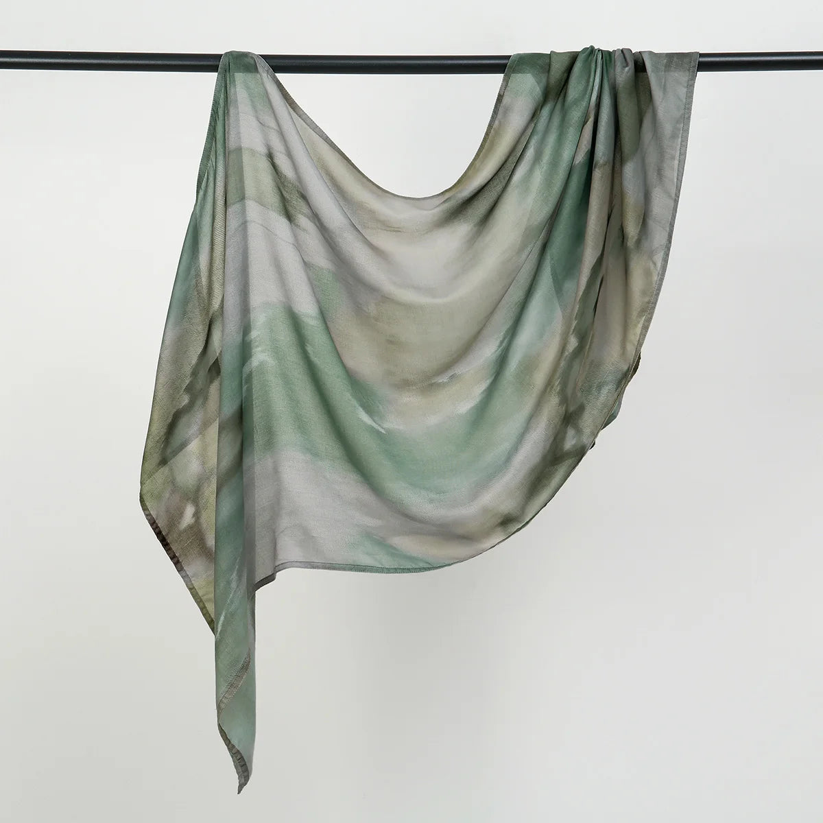 Durchscheinender, fließender Hijab-Schal in watercolorartigen grün-grauen Tönen an einem schwarzen Stäbchen aufgehängt.