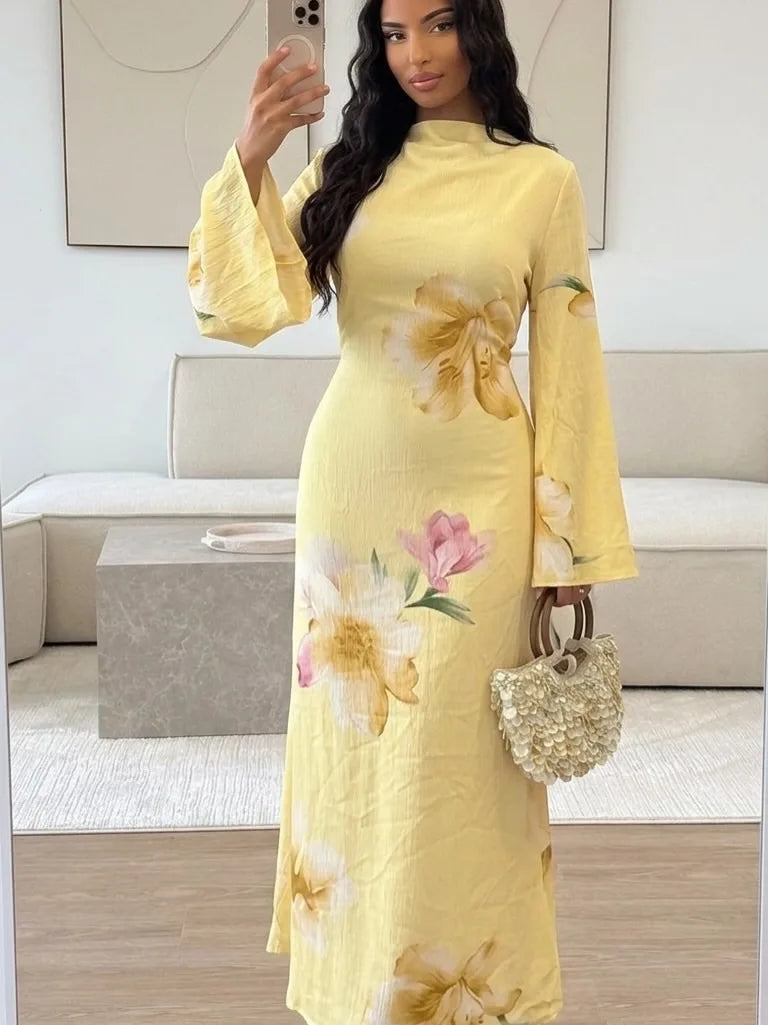 Eine Frau modelliert ein langes, gelbes, florales Abaya-Kleid mit langen Ärmeln und hohem Kragen in einem hellen Innenraum.
