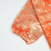 Close-up of an orange satin kaftan fabric with a subtle gold pattern and gathered cuff detail.