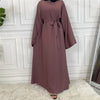 Elegane mauve maxi Abaya für Damen mit tiegter Taille und langen Ärmeln vor weißer Wand