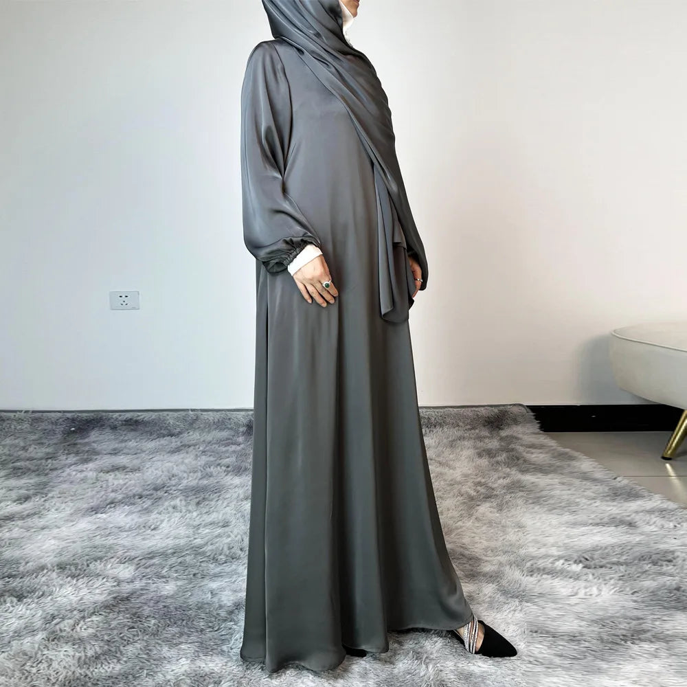 Una mujer vistiendo un largo y fluido vestido maxi de satén gris y hijab a juego, de pie en una habitación luminosa.