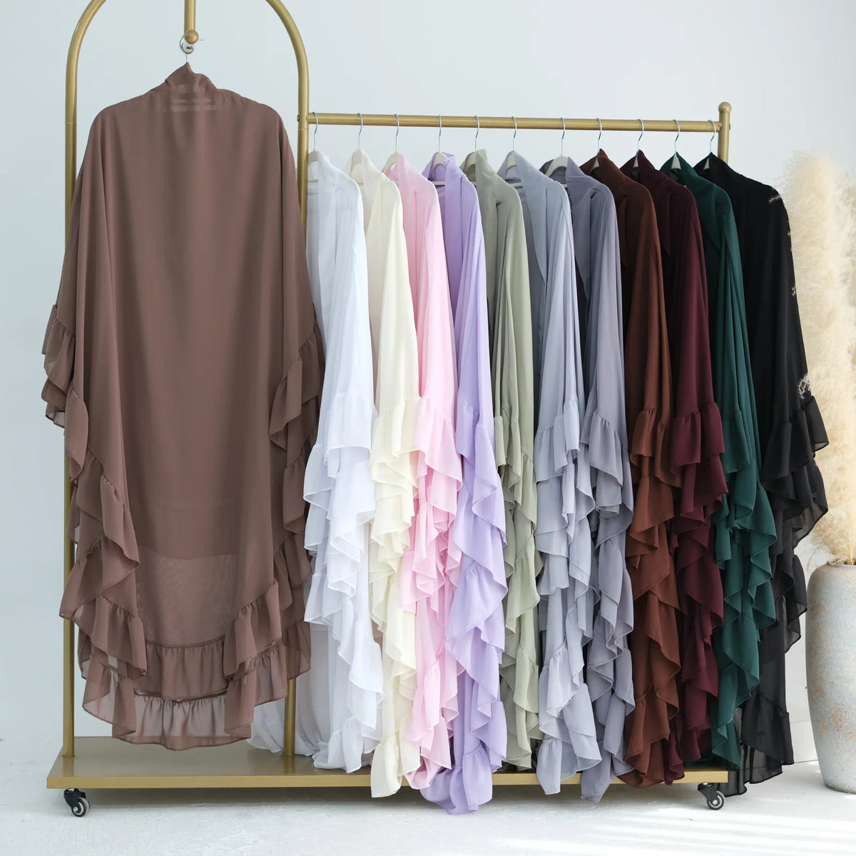 A display of various ruffled 2-layer chiffon hijabs in shades of pink, purple, green, and brown hanging on a gold rack.