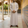 Un hombre vistiendo una jubba thobe bordada beige claro, robe casual, de pie en un pasillo con azulejos y columnas decorativa