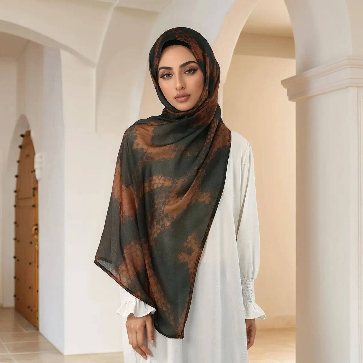 Eine Frau trägt einen eleganten bedruckten braun-schwarzen Hijab und ein weißes Kleid in einem hellen Flur.