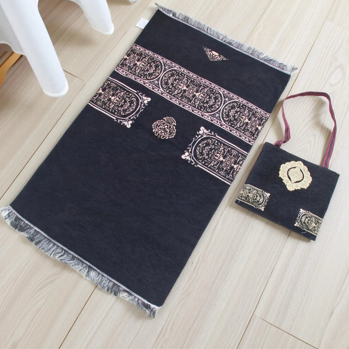 Dunkelgraue Gebetsmatte mit filigranen Blumenmustern und einer passenden kleinen Tasche auf einem hellen Holzfußboden.
