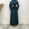 Eine Frau in einem eleganten dunkeltürkisen Maxi-Abaya mit langen Ärmeln und einem zugebundenen Bund vor einer weißen Wand.