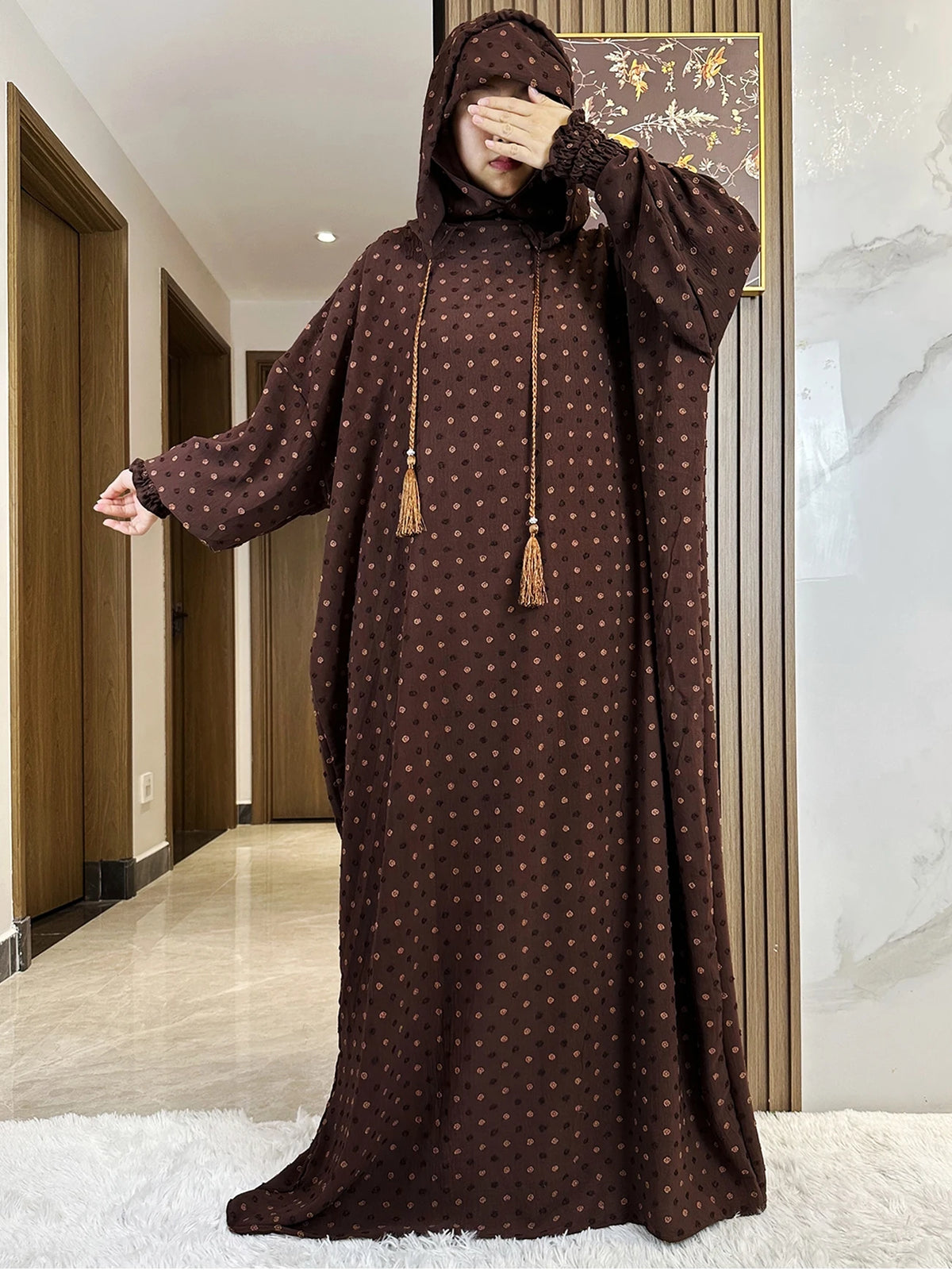 Una modelo vistiendo una abaya de algodón marrón oscuro con capucha y estampado de puntos, de pie en una habitación bien ilum