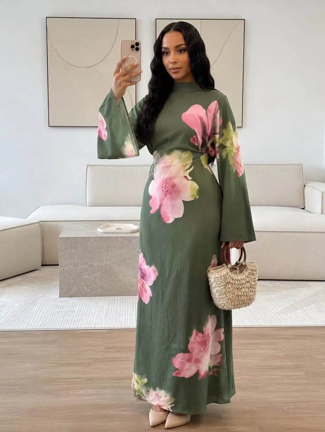 Eine Frau modelliert ein elegantes, langärmeliges olivgrünes Abaya-Kleid mit rosa Blumenmuster in einem hellen Wohnzimmer.