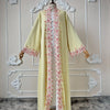 Une longue abaya jaune pâle avec bordure de dentelle florale rose, présentée sur un mannequin devant un fond arqué blanc.