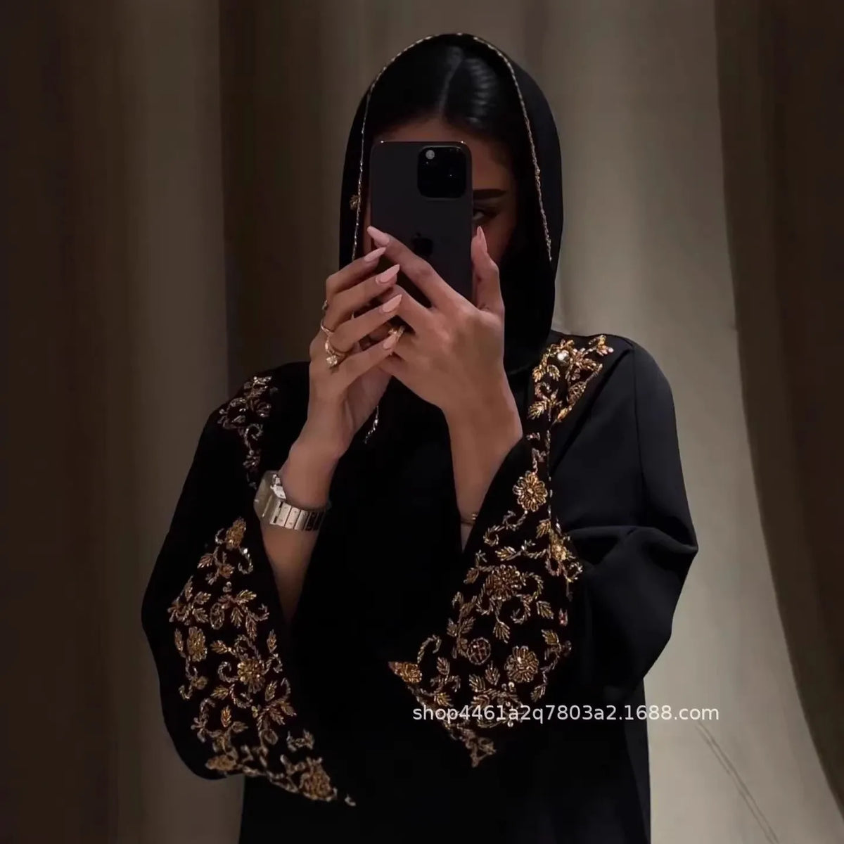 A woman wearing a black abaya with gold floral beadwork, holding a smartphone.