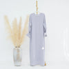 Un largo vestido abaya lavanda claro con puños fruncidos, exhibido en un perchero junto a un jarrón de pampas seco beige.