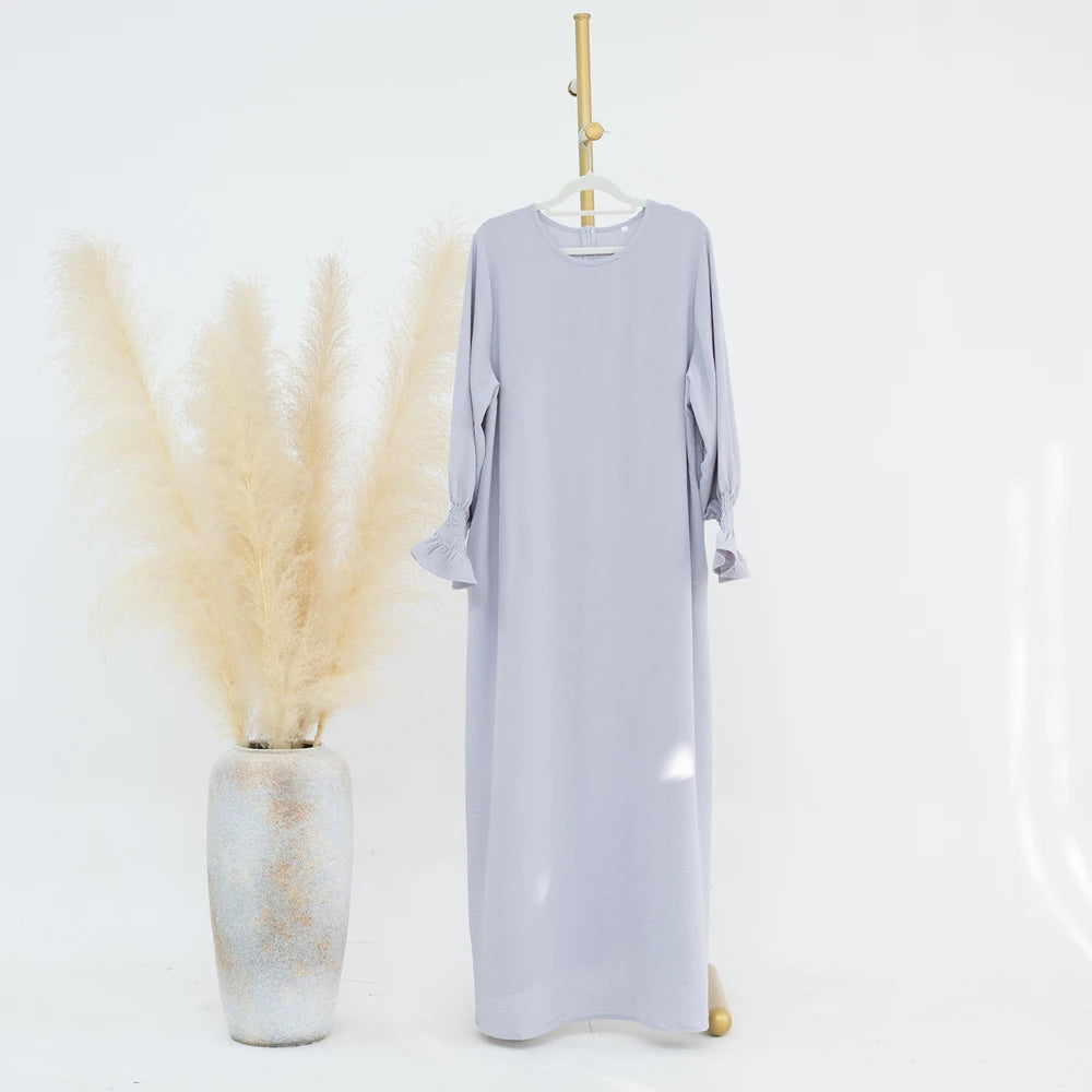 Un largo vestido abaya lavanda claro con puños fruncidos, exhibido en un perchero junto a un jarrón de pampas seco beige.