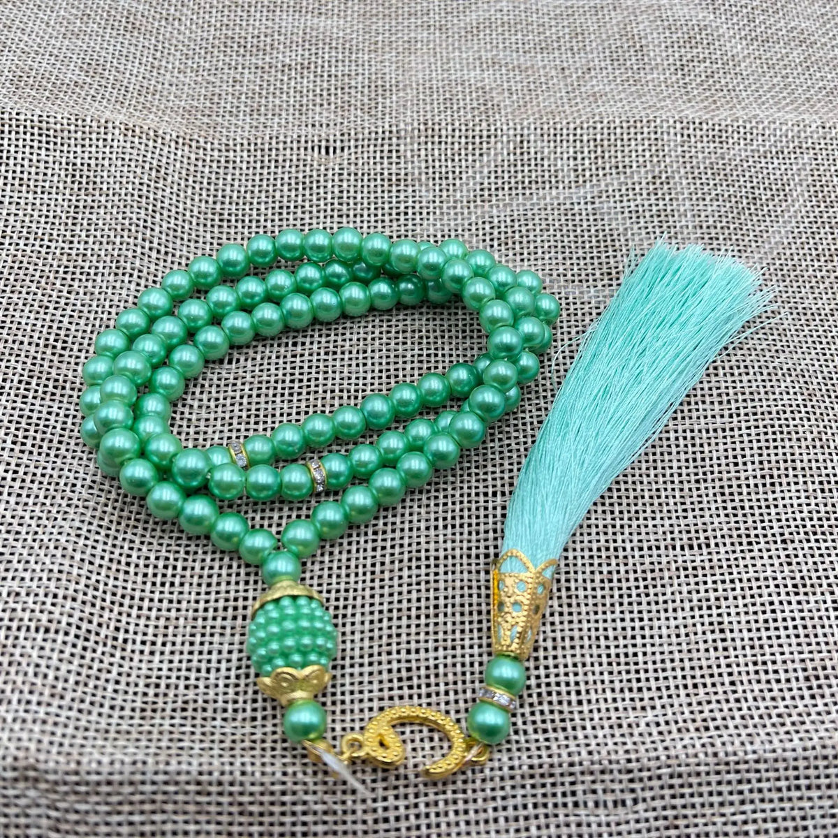 Grüne Perlenrosary-Armband mit türkisem Quaste auf beigem Stoffhintergrund