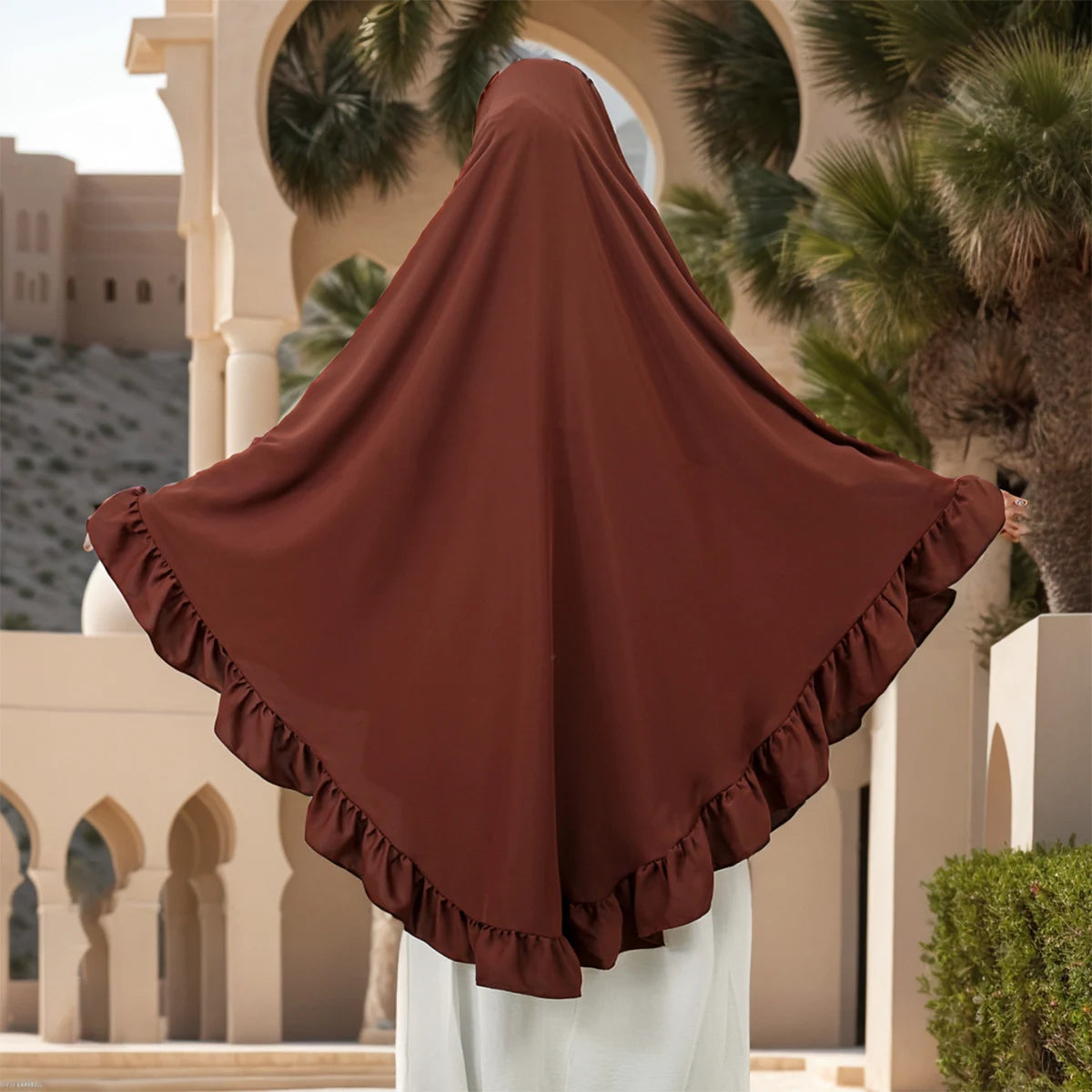 Drapierte, rostfarbene muslimische Hijab-Schal mit gerafften Rändern vor architektonischer Kulisse und Palmen.
